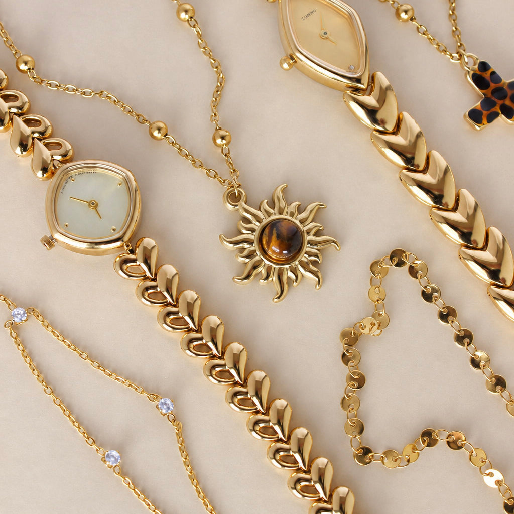 A collection of gold-toned jewelry and watch accessories arranged on a light beige background, including bracelets, chains, a necklace with a sun pendant, and watch parts.