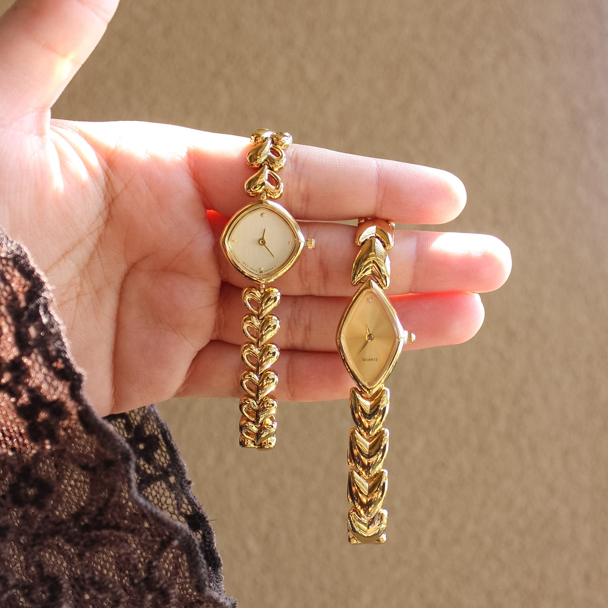 A pair of gold-toned watches set held in a person's hand against a warm beige background.