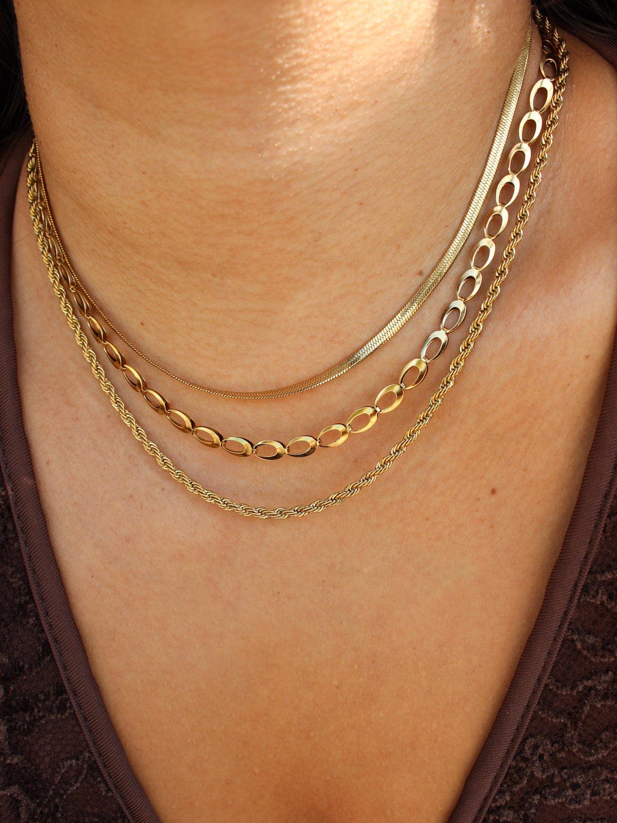 A close-up of a person's neck and upper chest wearing three layered gold necklaces: a thin curved chain, a curb chain, and a delicate rope chain.