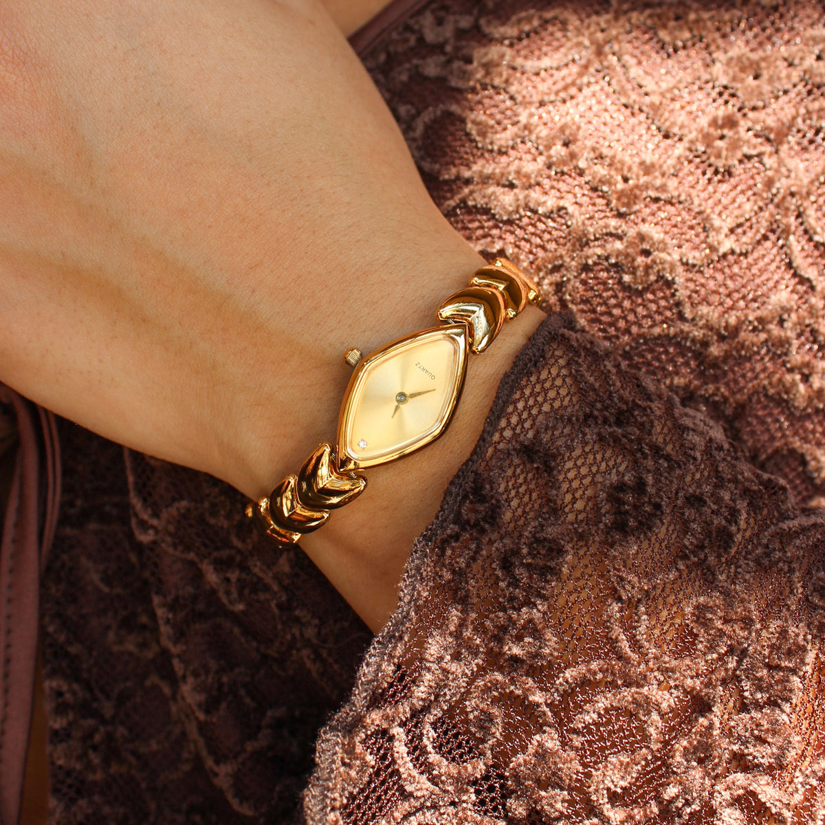 A close-up of a woman's wrist wearing a gold-tone watch with a diamond-shaped dial and matching bracelet, styled with a brown lace top.