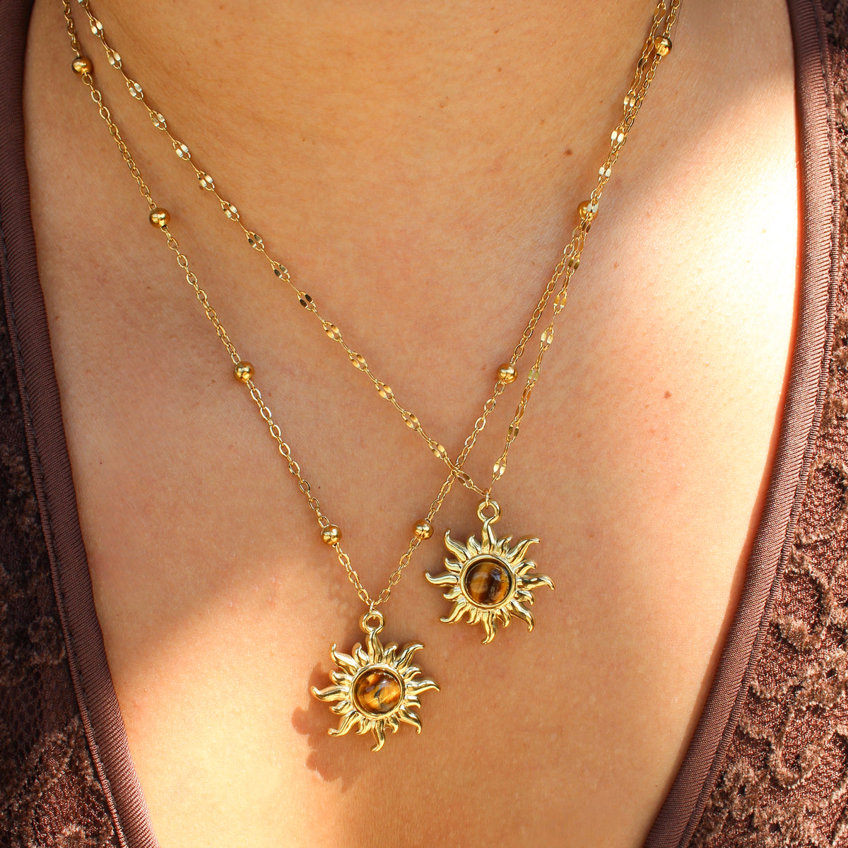 A close-up of a person wearing two layered gold necklaces with sunburst pendants and amber-colored round stones, styled with a brown lace top.