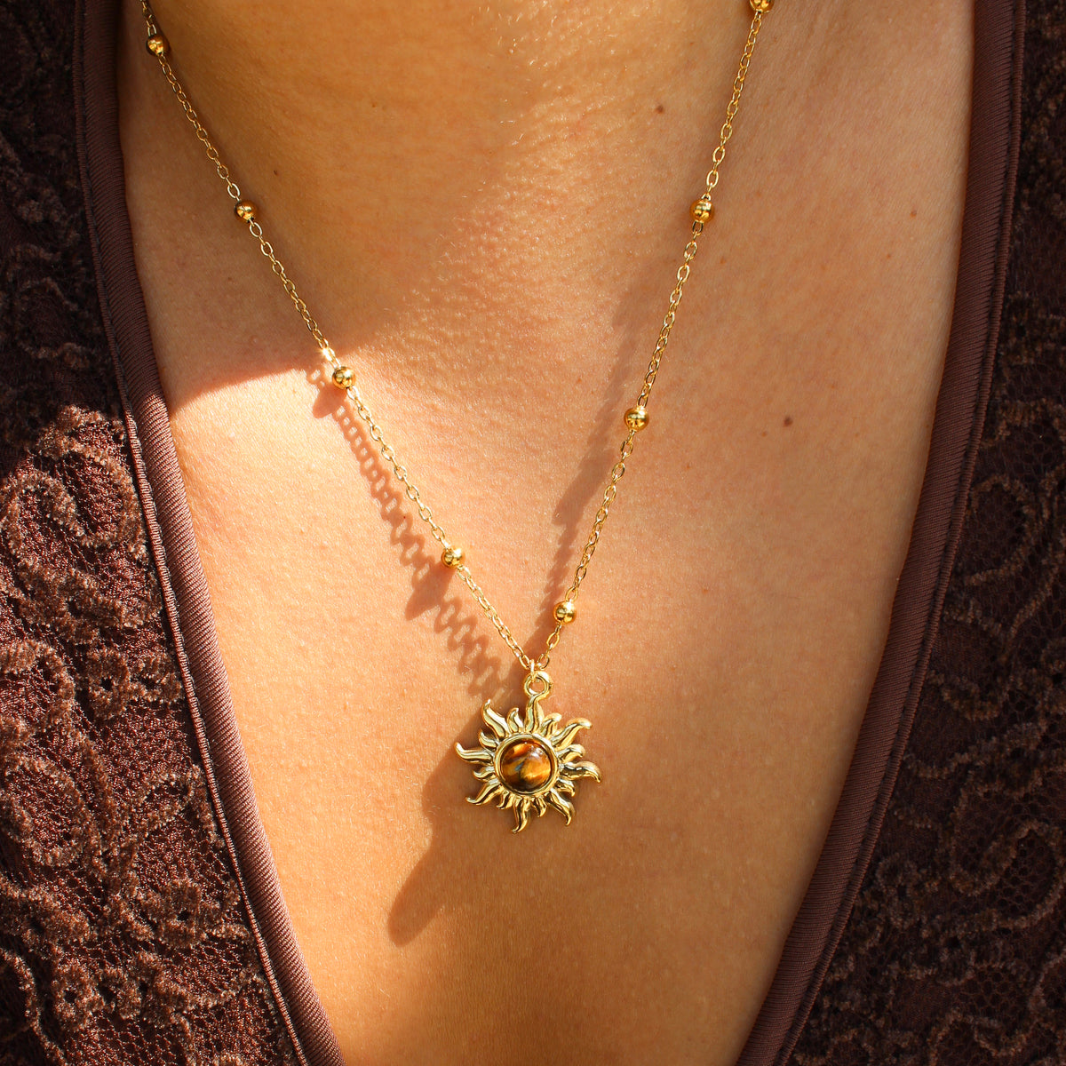 A gold necklace with a sun pendant and small bead accents, shown worn on a person's neck over a brown lace top.