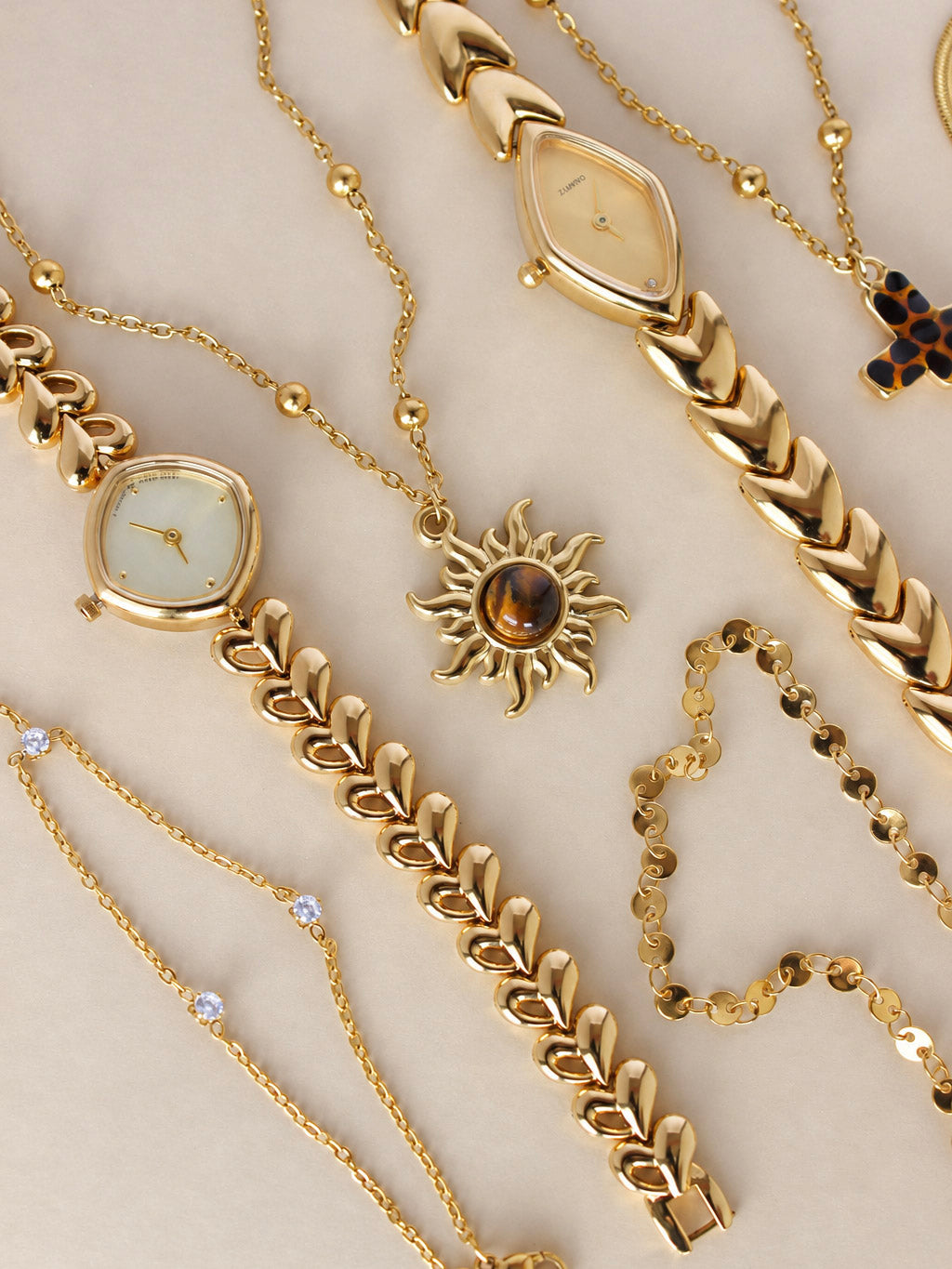 A collection of gold-toned jewelry and watch accessories arranged on a light beige background, including bracelets, chains, a necklace with a sun pendant, and watch parts.