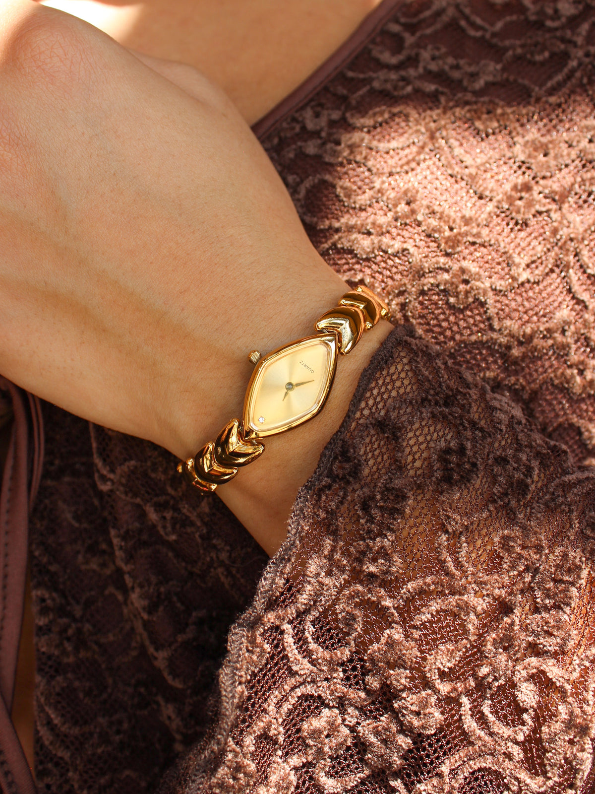 A close-up of a woman's wrist wearing a gold-tone watch with a diamond-shaped dial and matching bracelet, styled with a brown lace top.
