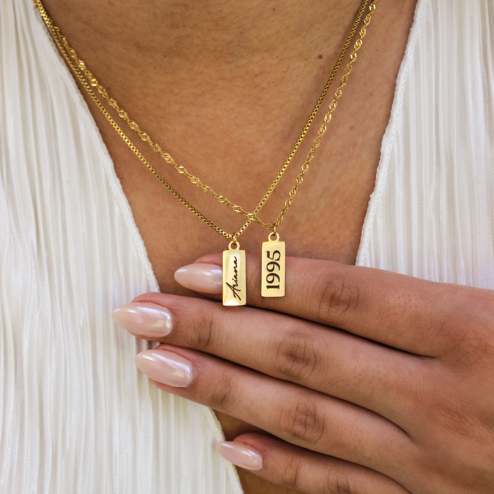 Gold necklaces with 'Ariana' and '1995' pendants on a person wearing a white top.