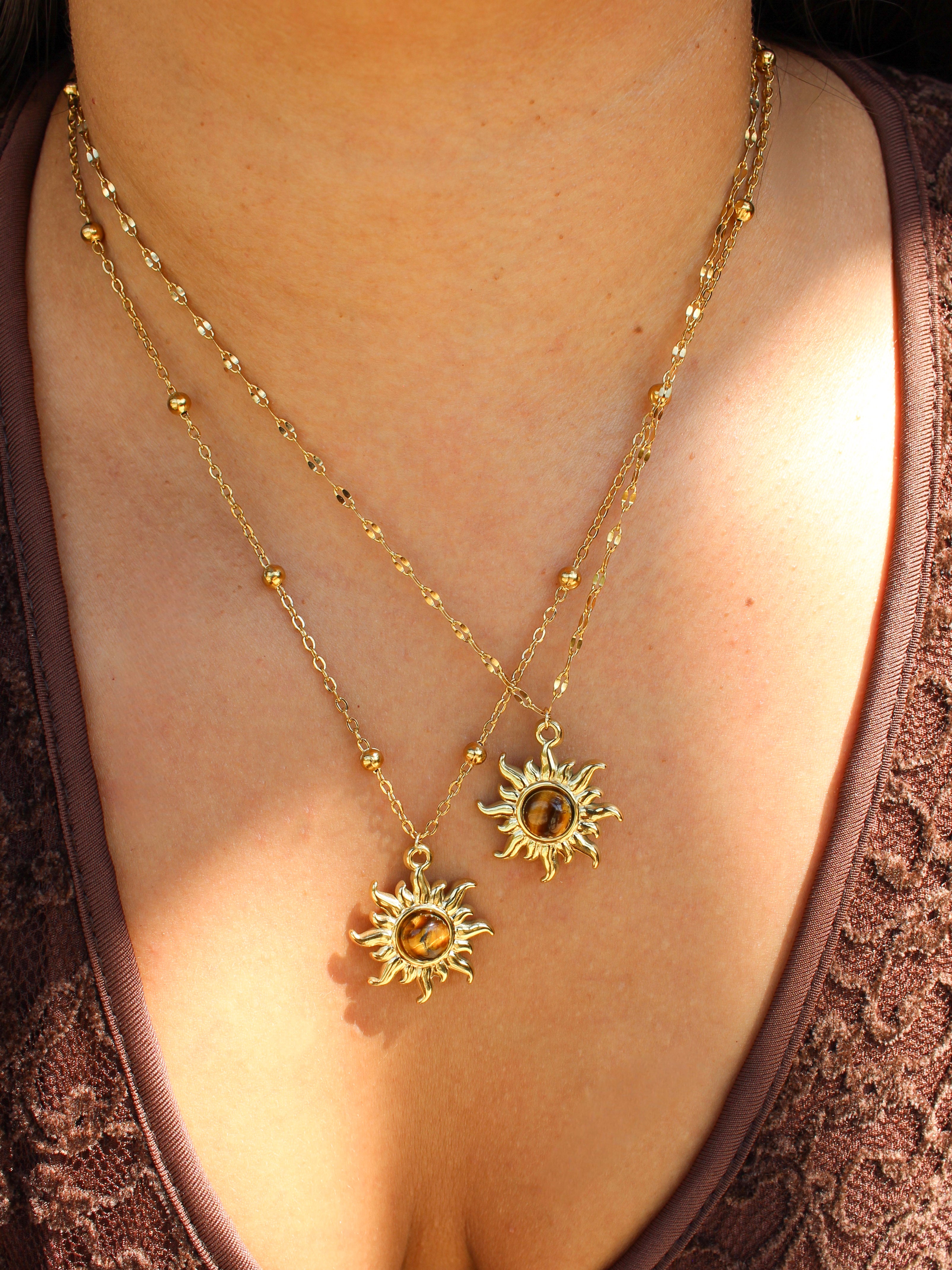 A close-up of a person wearing two layered gold necklaces with sunburst pendants and amber-colored round stones, styled with a brown lace top.