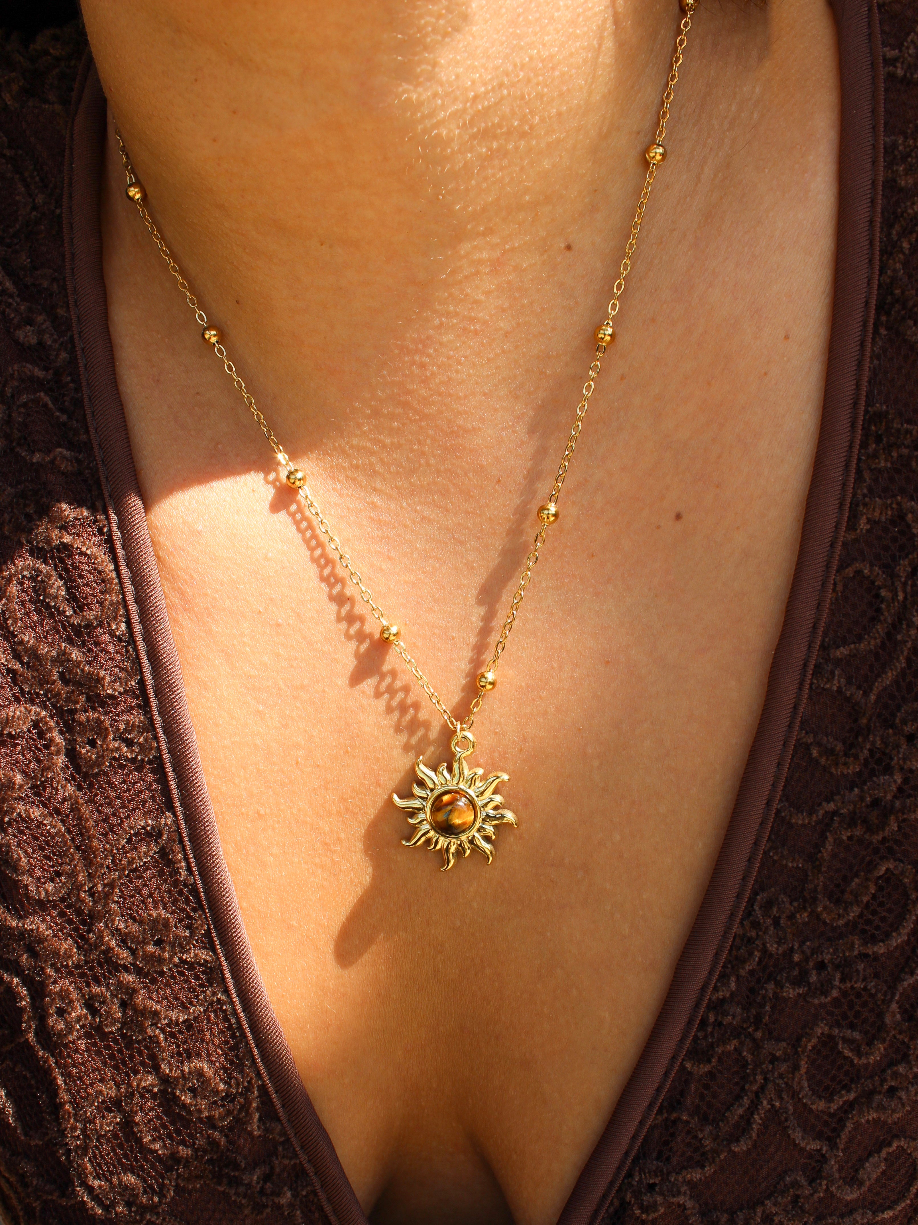 A gold necklace with a sun pendant and small bead accents, shown worn on a person's neck over a brown lace top.